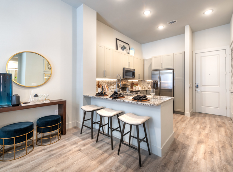 Staged kitchen with breakfast bar, granite countertops, white fabric topped barstools, stainless appliances, taupe cabinetry, circular mirror, wood style flooring