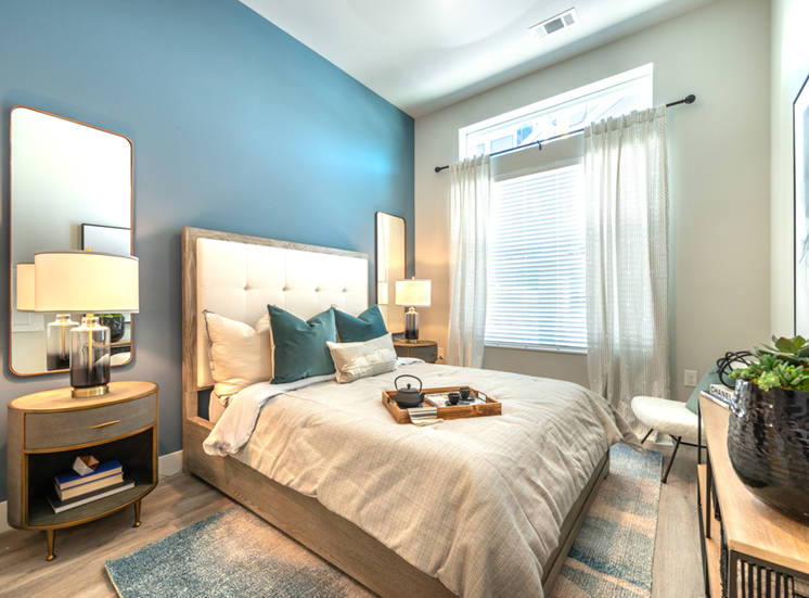 Staged bedroom with bed, accent rug, wood style flooring, oval side tables with lamps and mirrors, large windows with blinds and sheer curtain with blue accent wall