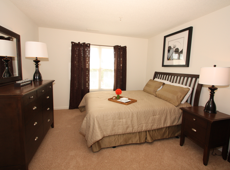 The furnished mater bedroom is carpeted throughout with a large picture window on one of the walls. Featured here is a queen size bed with tan comforter and coordinating pillows, espresso colored dresser and nightstand, and a single lamp on top of each.