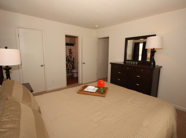 The furnished master bedroom is carpeted throughout with direct access to the bathroom. There is a queen size bed with tan comforter and matching pillows, a large dark wooden dresser with a lamp and mirror, and a matching nightstand with a lamp on top.