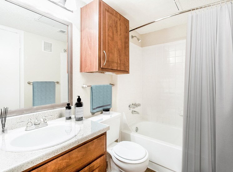 Staged bathroom with tiled bathtub shower combo. White counter tops and brown cabinets with wood style flooring.