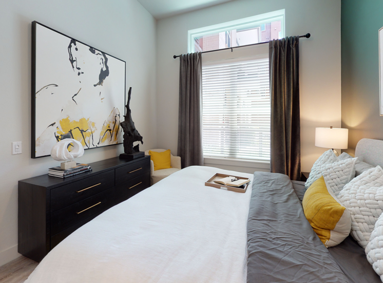 Staged bedroom with bed, dresser, art on wall, accent wall, large window with blinds and curtain