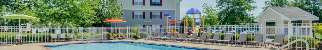 The community pool is rectangular in shape and is surrounded by a spacious sundeck with taupe pool lounge chairs, and two round picnic tables with umbrellas in the middle of the table. The pool is enclosed by a white iron fence. Outside of the pool area is the community playground, the mailbox station, apartment buildings, and mature trees.