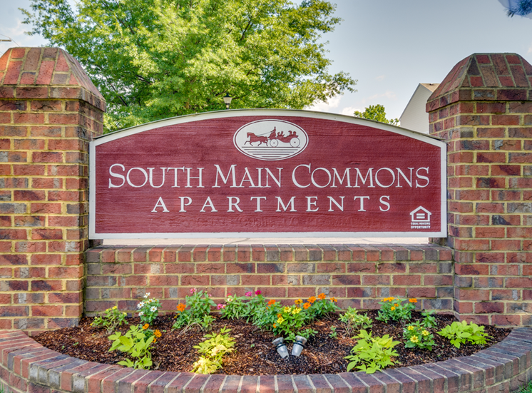 The main sign by the entrance is made of brick, with two columns on either side. In between the columns is a red wooden sign with the community name and logo on it. There is a small flower bed at the base of the sign.