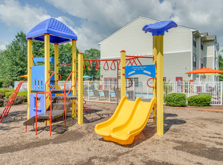 The community playground is red, yellow and blue. It has a ladder, monkey bars, stairs, and two yellow slides. The playground area has mulch on the ground and is located near apartment buildings and the community pool.
