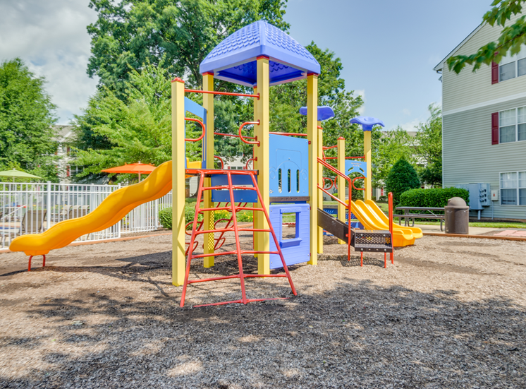The community playground is red, yellow and blue. It has a ladder, monkey bars, stairs, and three yellow slides. The playground area has mulch on the ground and is located near the pool and apartment buildings.
