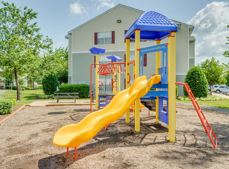 The community playground is red, yellow and blue. It has a ladder, monkey bars, stairs, and a yellow slide. The playground area has mulch on the ground and is located near apartment buildings and parking.