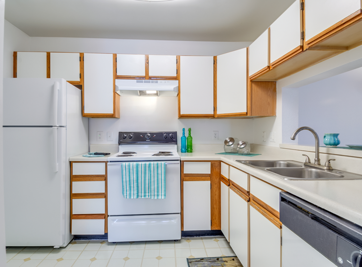 The three bedroom kitchen has white cabinets with tan trim, white appliances, formica countertops, and a double basin sink with gooseneck faucet. The breakfast bar has a place setting of fiesta ware for two.