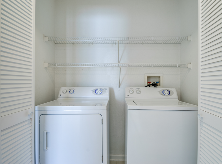 The laundry room is enclosed by two accordion, white, wooden doors. There is a washer and dryer with built-in shelving above them.