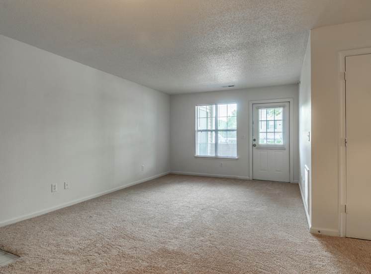 The vacant apartment living room has wall to wall carpet white walls, large picture window and a door leading out to the patio.