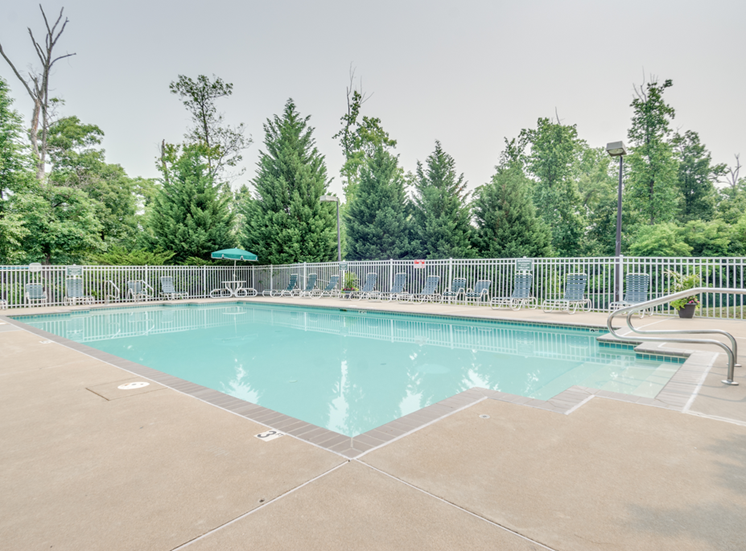 The community pool is rectangular in shape with blue and green pool furniture placed around the sundeck. There is a round picnic table with a green umbrella in the middle. The pool is placed on the back side of the Leasing Office.