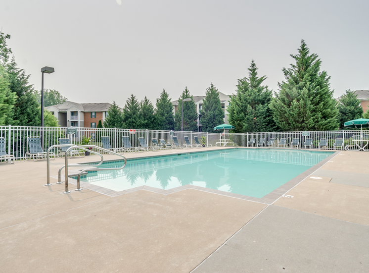 The community pool is rectangular in shape with blue and green pool furniture placed around the sundeck. There is a round picnic table with a green umbrella in the middle. The pool is placed on the back side of the Leasing Office.