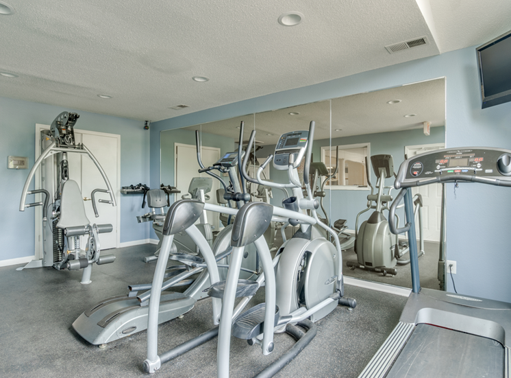 The community fitness center is equipped with a treadmill, two ellipticals and a weightlifting machine. One walls is made of mirrors and there is a TV mounted in the corner.
