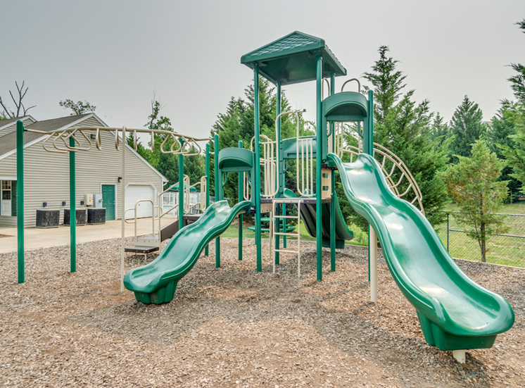 The community playground is surrounded by mulch and mature trees, and is located to the side of the clubhouse. The jungle gym is green and tan, has stairs leading to three slides and monkey bars.