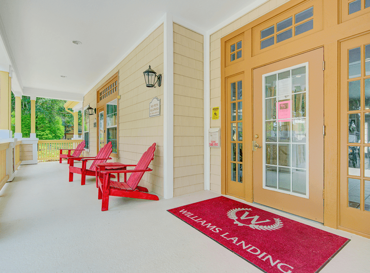 Exterior clubhouse entrance with covered porch featuring red adirondack chairs, red welcome mat, and tan door frame entrance