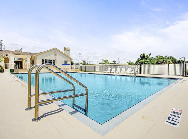 Swimming pool and sundeck with tables, chairs and loungers. The perimeter fence is lined with trees and the exterior of the clubhouse.
