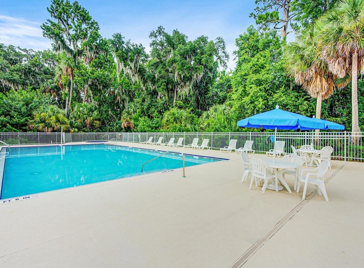 Swimming pool and sundeck with tables, chairs, and loungers. The perimeter fence is lined with trees.