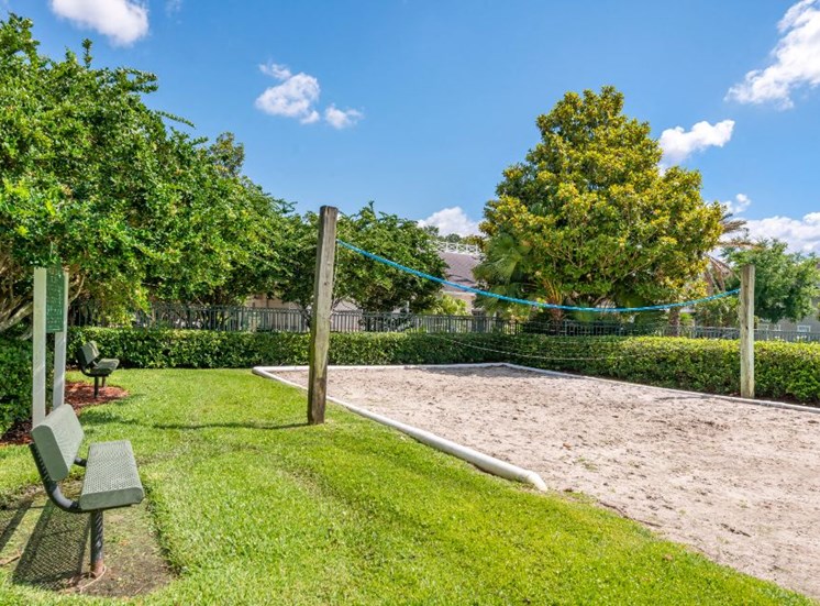 Sand Volleyball Court with Nearby Bench Surrounded by Trees and Shrubs with Building Exterior in The Background