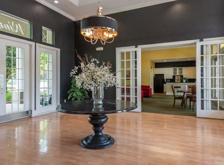 Clubhouse Foyer with a Table in the Middle with A Vase and Flowers on it Hardwood Floors Large Windows and Open Doors Leading to Clubhouse Seating Area with Kitchen