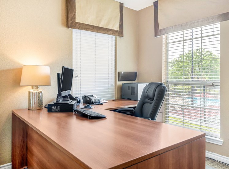 Leasing desk with monitor, keyboard, mouse, phone, lamp, and printer on top with chair and 2 windows in the background