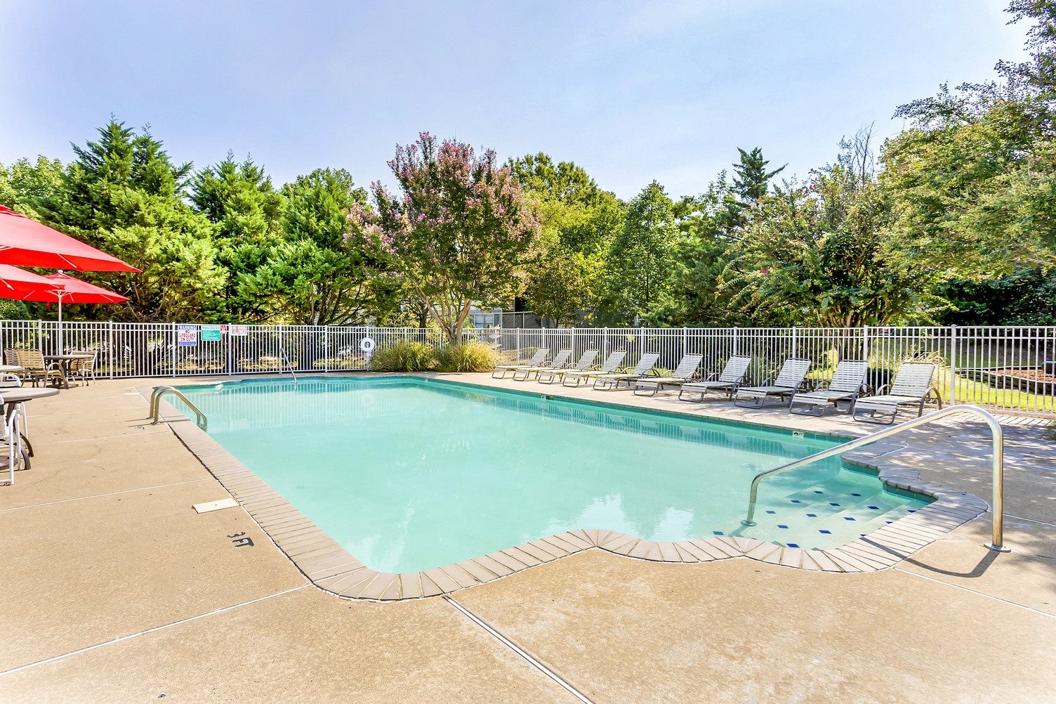 Community pool with sundeck, lounge chairs, surrounded by white metal fence with trees in the background