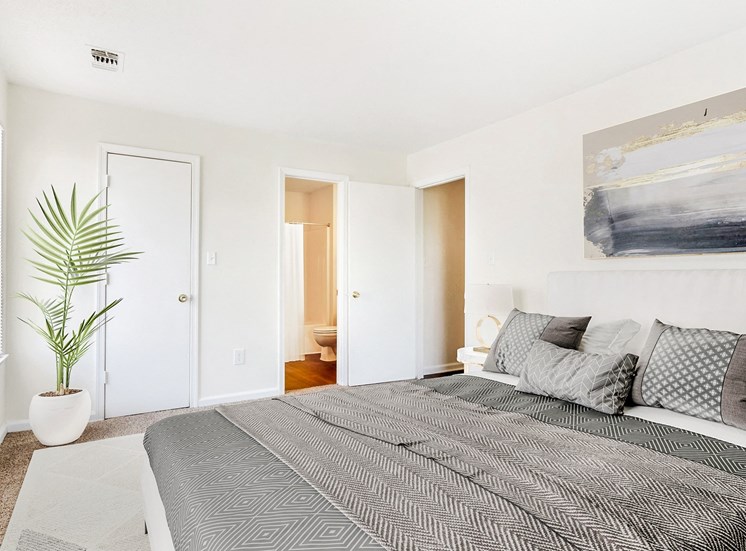 Staged bedroom with queen sized bed, art above bed, nightstand, large window in the background and view of bathroom and hall.