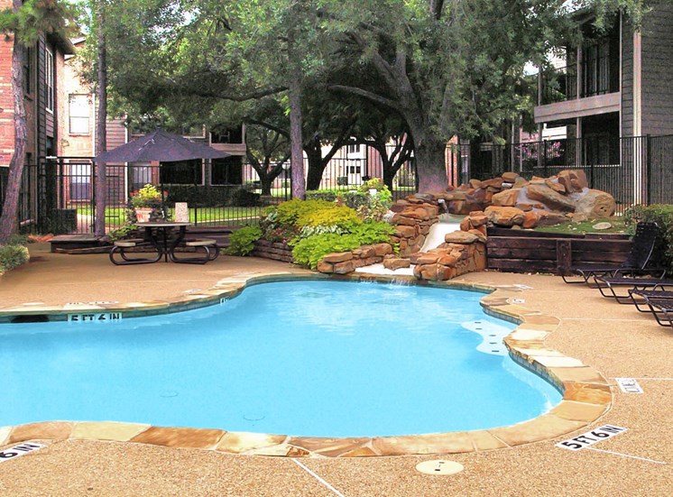 Swimming pool with stone waterfall surrounded by sundeck, green landscaping, picnic table with umbrella and building exteriors in the background