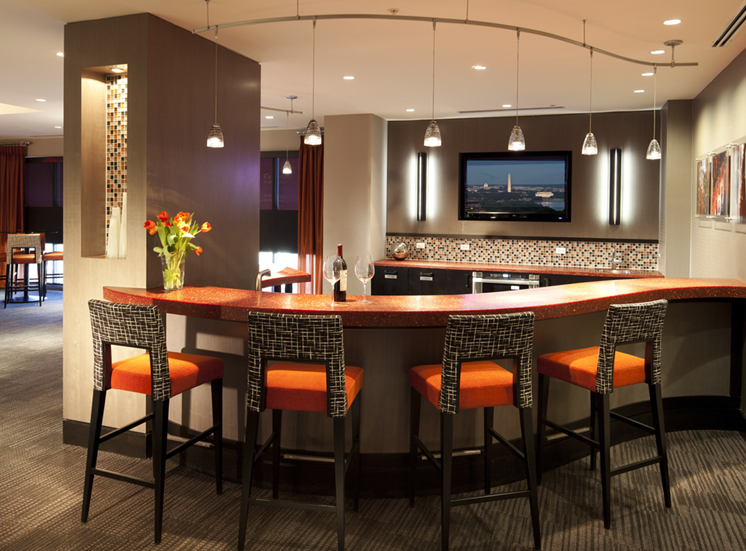 Clubroom kitchen. Bar area with four bar stools overlooking kitchen with tv on wall. Orange and wood accent colors with artwork on wall. Additional seating in background, 4 top table.