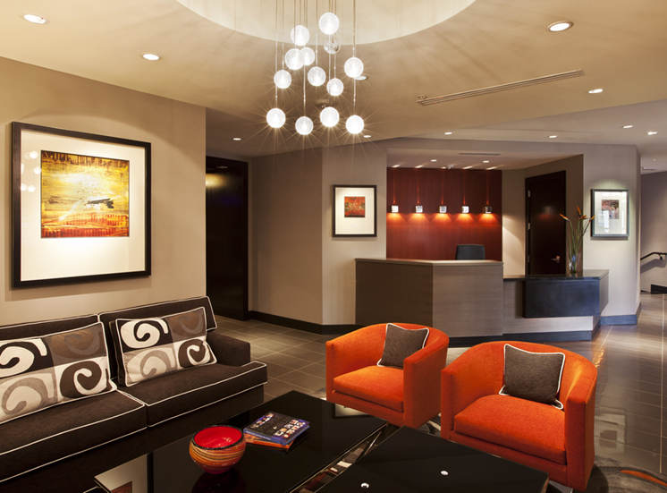 Clubroom seating with couch and two orange accent chairs with coffee table. Leasing desk and office in background.
