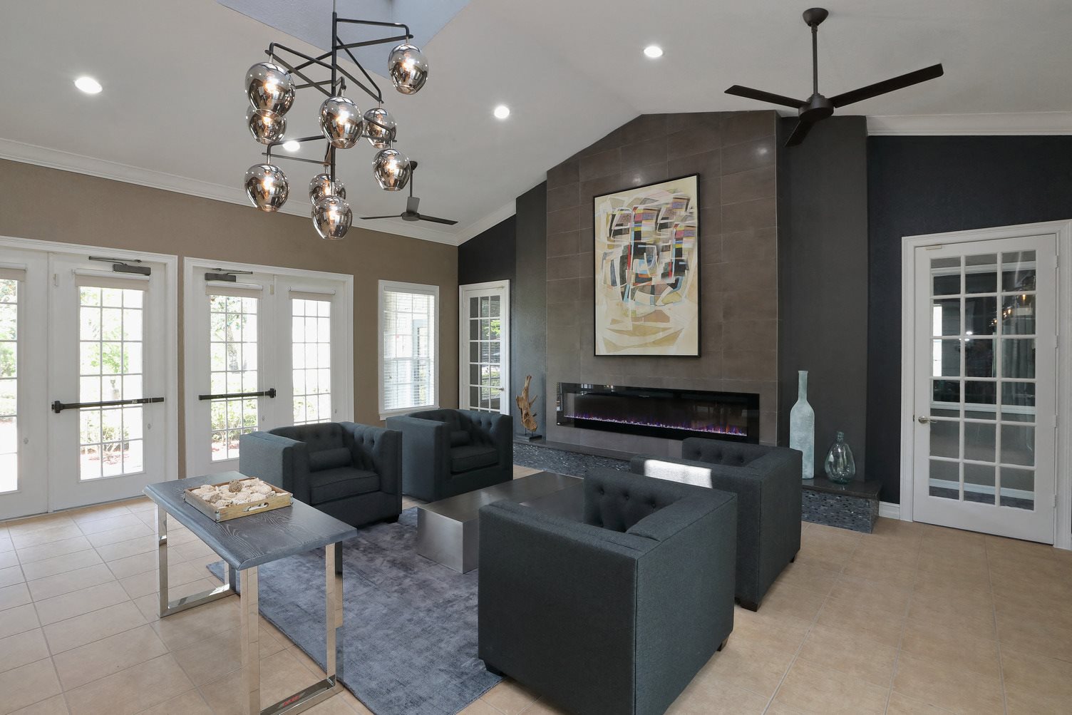The Resident Lounge is a big room with a contemporary design. The far wall shows a large painting artwork above a fireplace. Four dark chairs are sat around a small table in front of french doors.