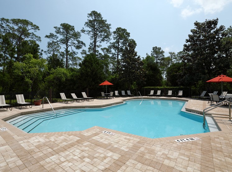 The swimming pool is a large area with calm turquoise waters with a brick paver sundeck surrounding. Light lounge chairs and orange umbrellas are placed around the sundeck.