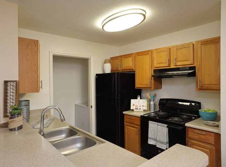 The B1 Kitchen has light oatmeal-colored countertops with light wood cabinetry. Black kitchen appliances contrast against the light paint. An open closet that at the end of the galley-style room holds the full-size washer and dryer appliances.