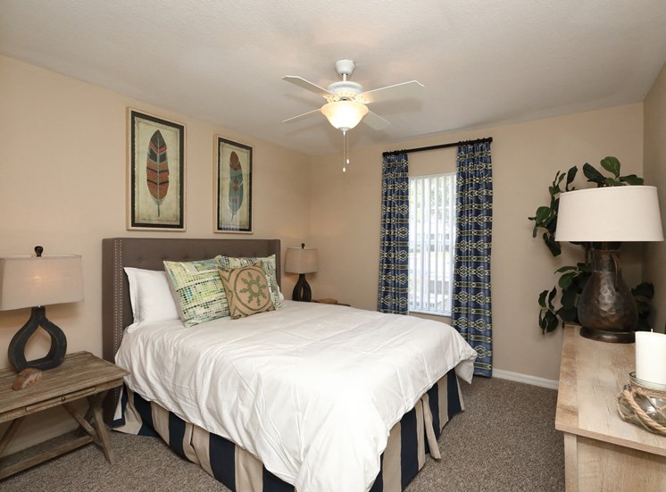 The Bedroom is a comfortable space with a large bed centered on the main wall. A large window brings plenty of natural light into the room. The carpeted flooring continues throughout with furniture filling spaces around the room.