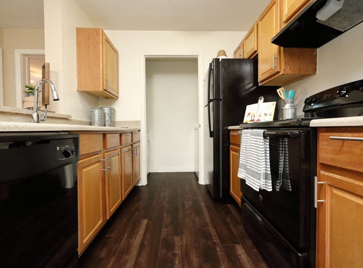 The B1 Kitchen has light oatmeal-colored countertops with light wood cabinetry. Black kitchen appliances contrast against the light paint. An open closet at the end of the galley-style room holds the full-size washer and dryer appliances.