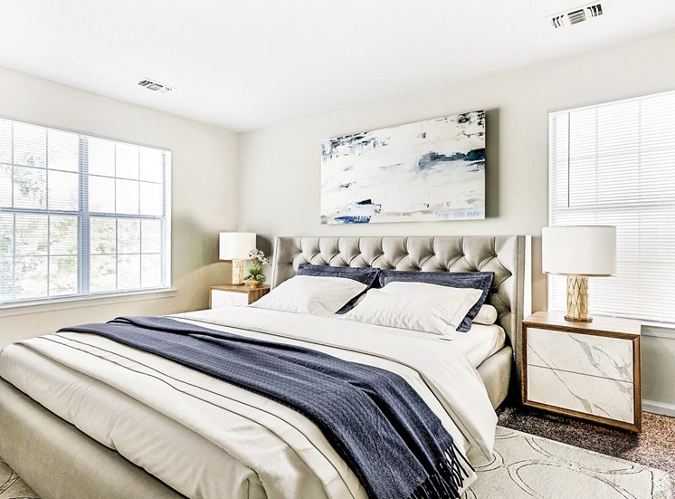 Staged bedroom with queen sized bed, art above bed, two nightstands, and large windows in the background.