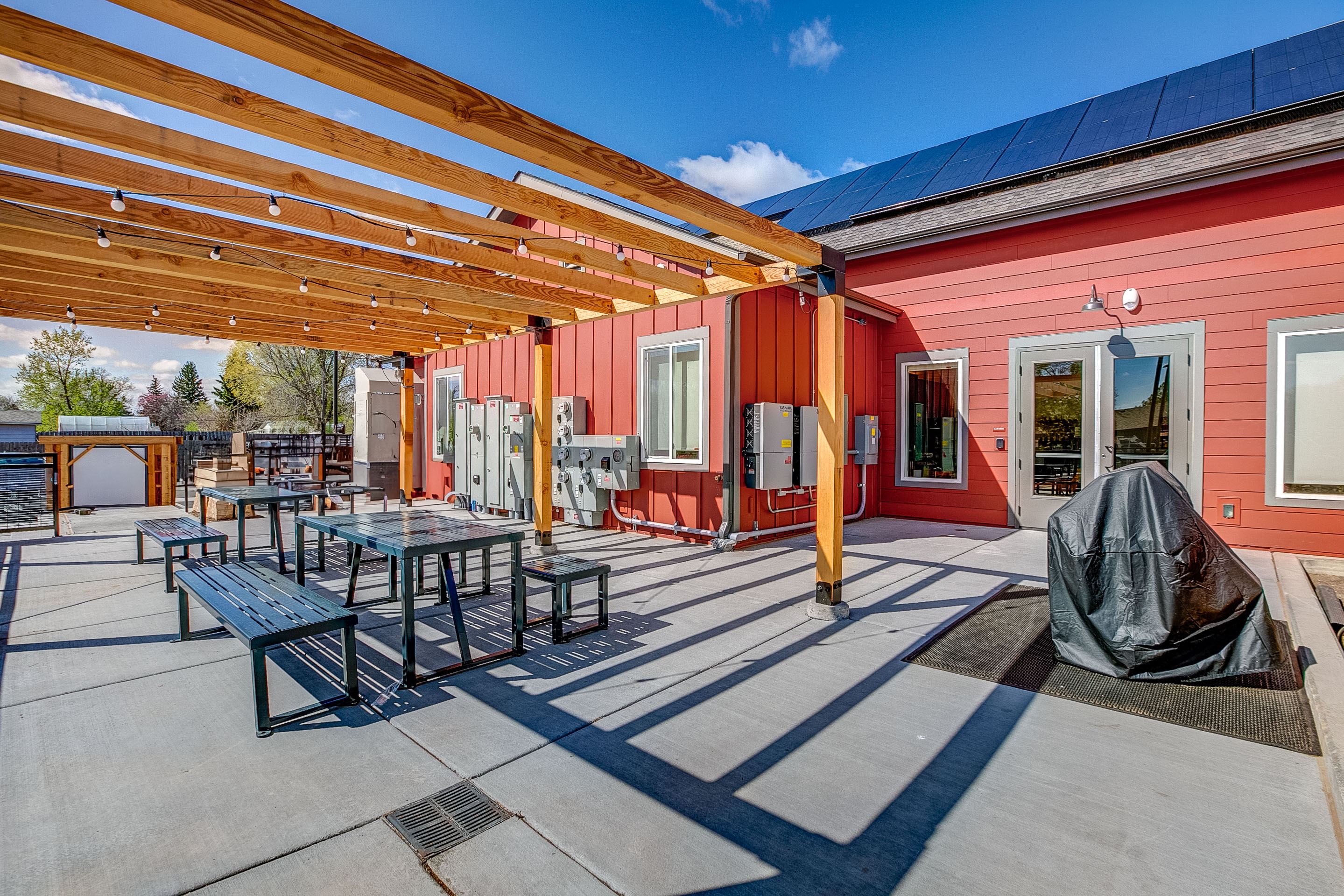 A red building with a wooden pergola over a patio with tables and chairs.