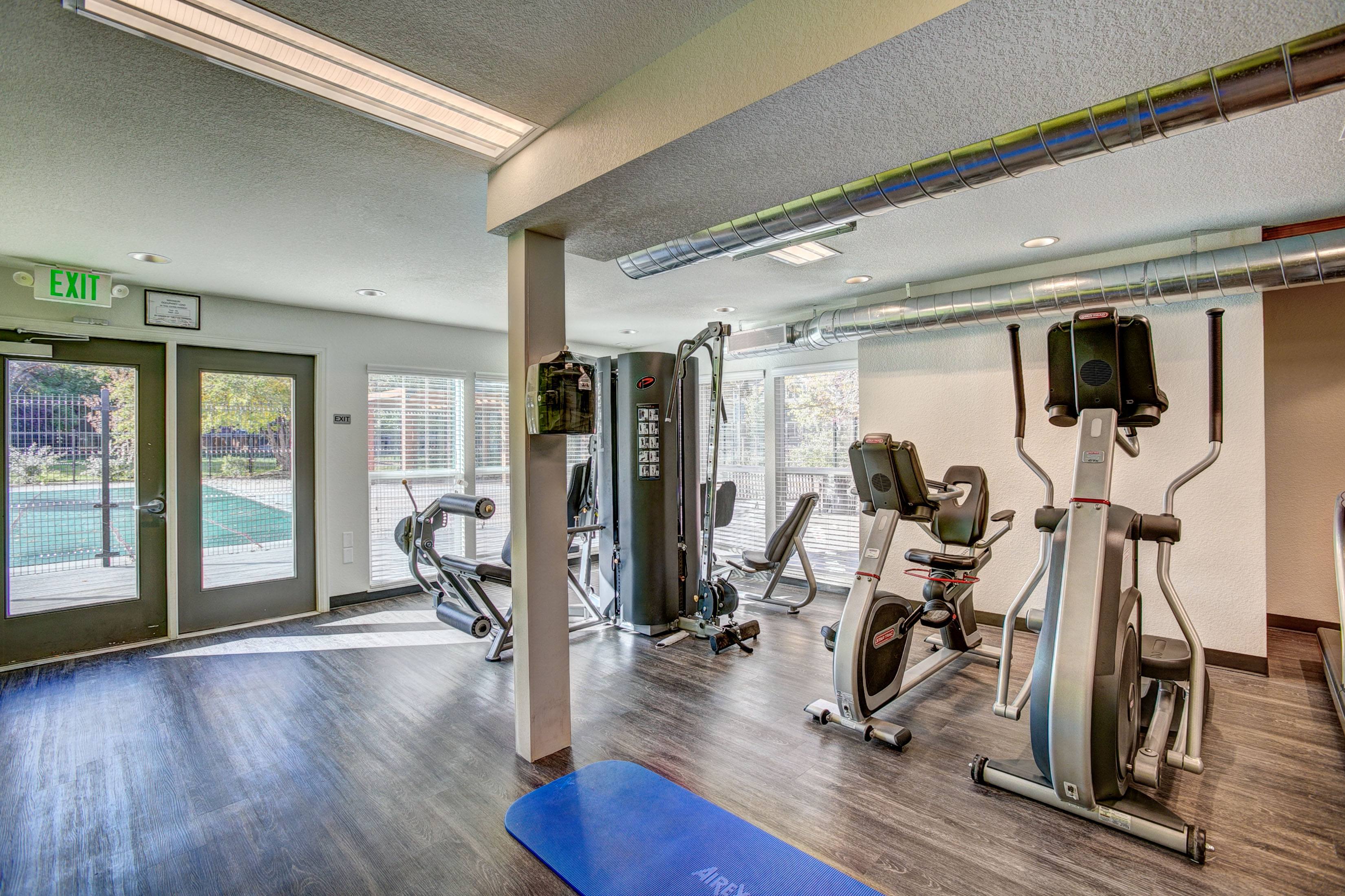 A gym with a blue mat in the foreground and a green exit sign on the wall.