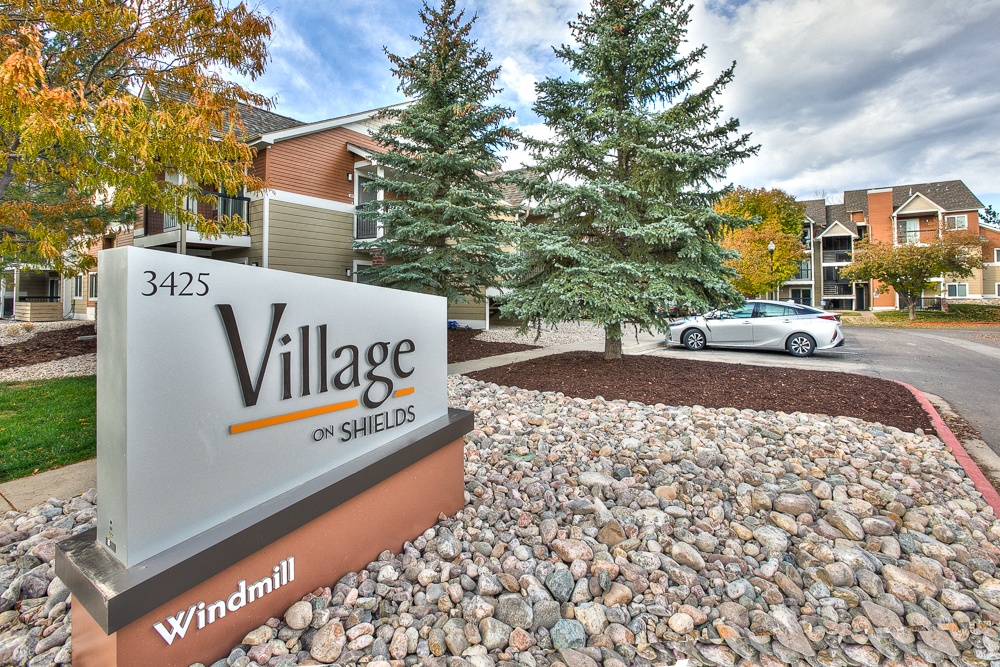A sign for Village on Shields stands in front of a gravel area.