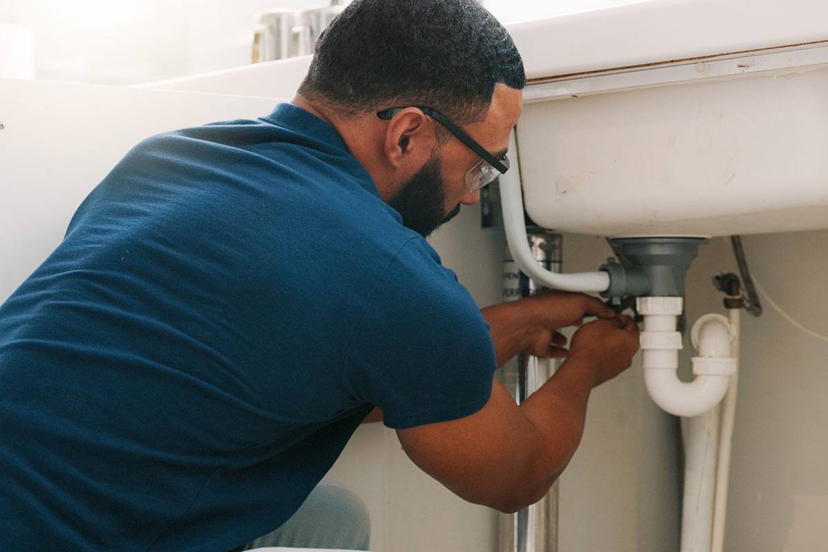 A man in a blue shirt is fixing a white pipe under a sink