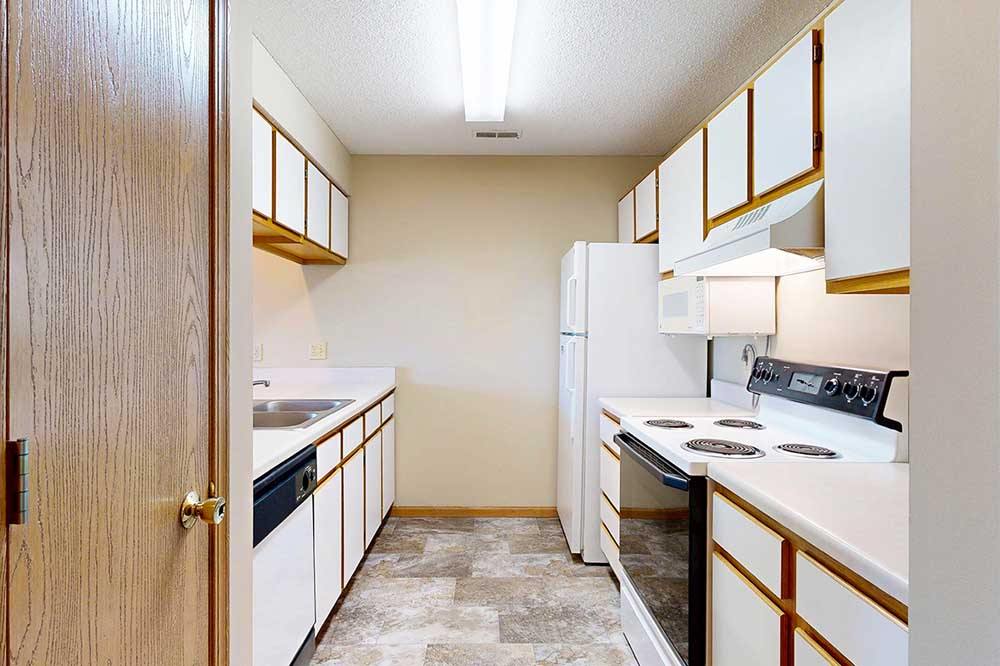 Galley style kitchen with white cabinets and oak trim, white appliances, and tan vinyl flooring