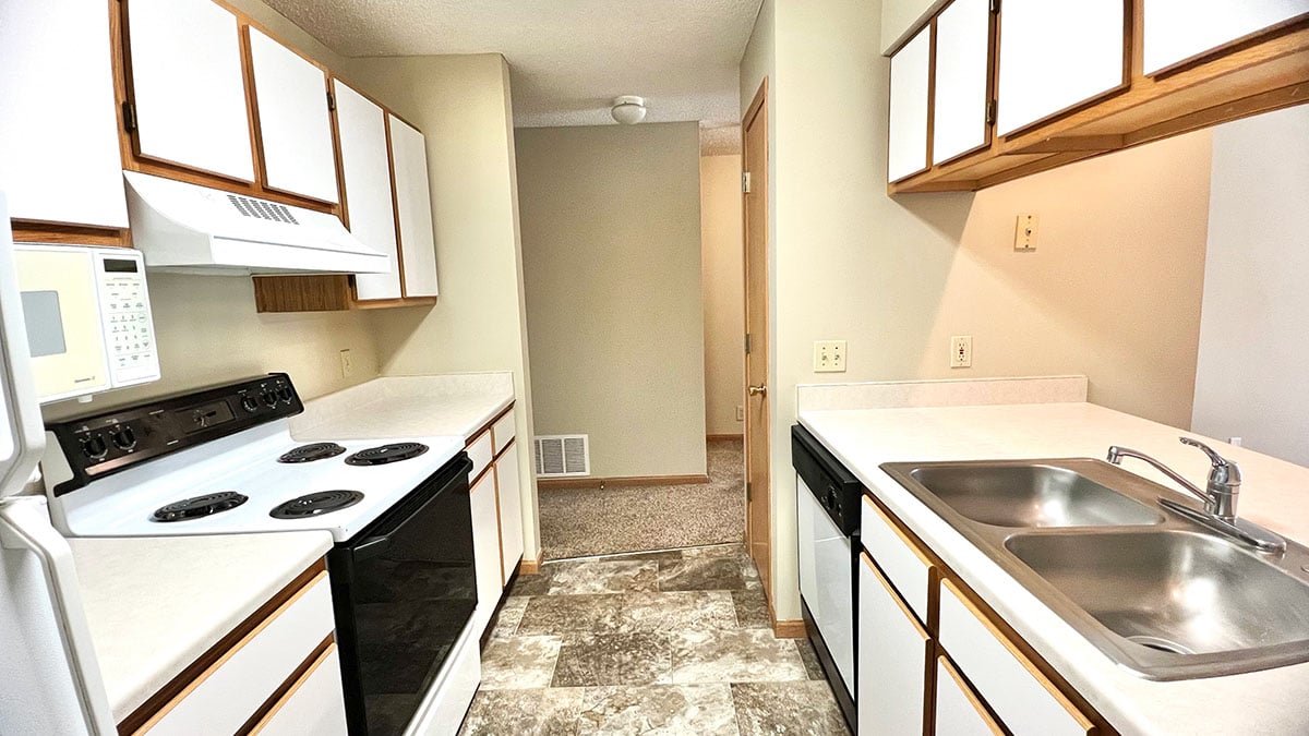 Galley style kitchen with white cabinets and oak trim, white appliances, and tan vinyl flooring