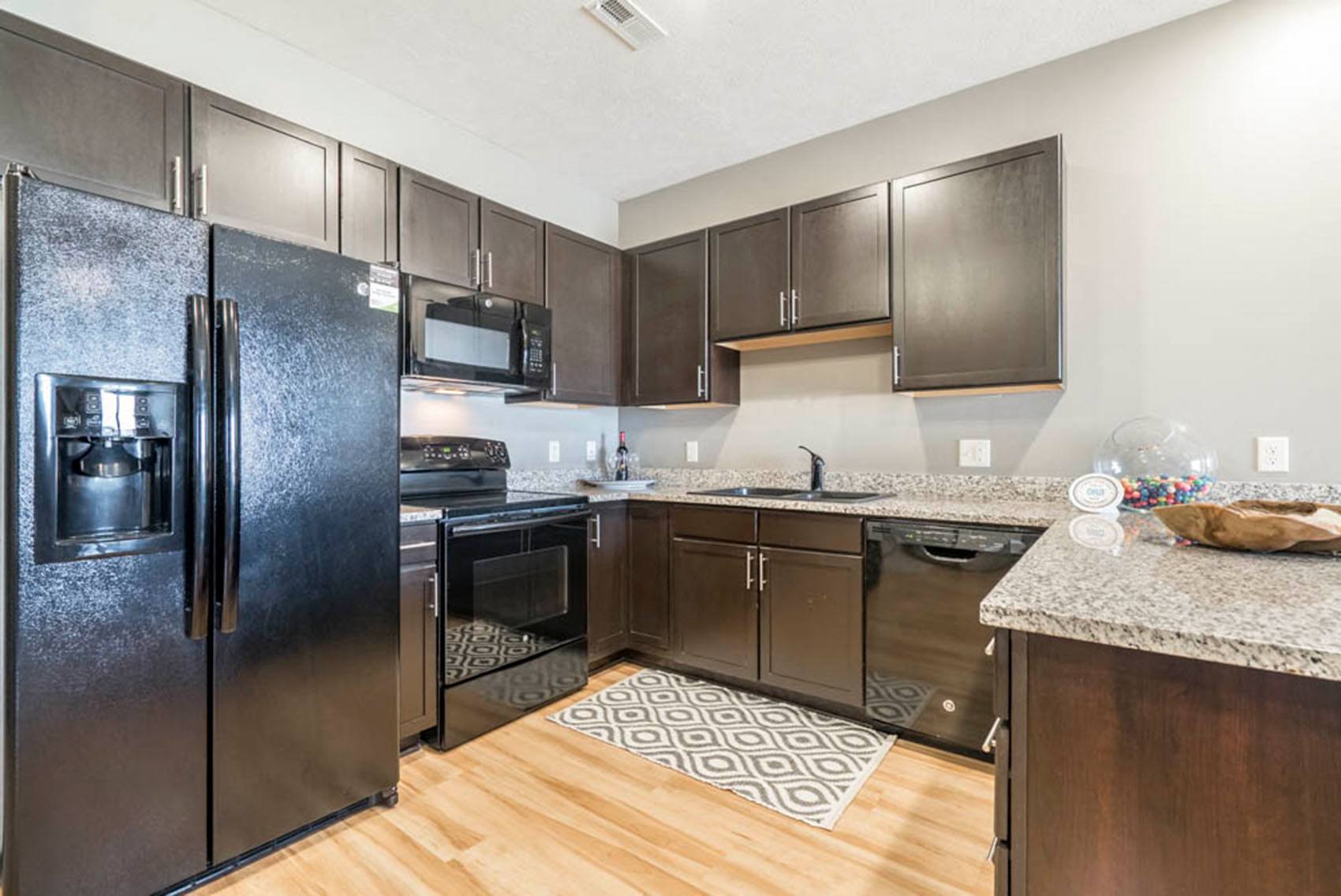 A modern apartment kitchen with black appliances, deep brown cabinets, and granite counters