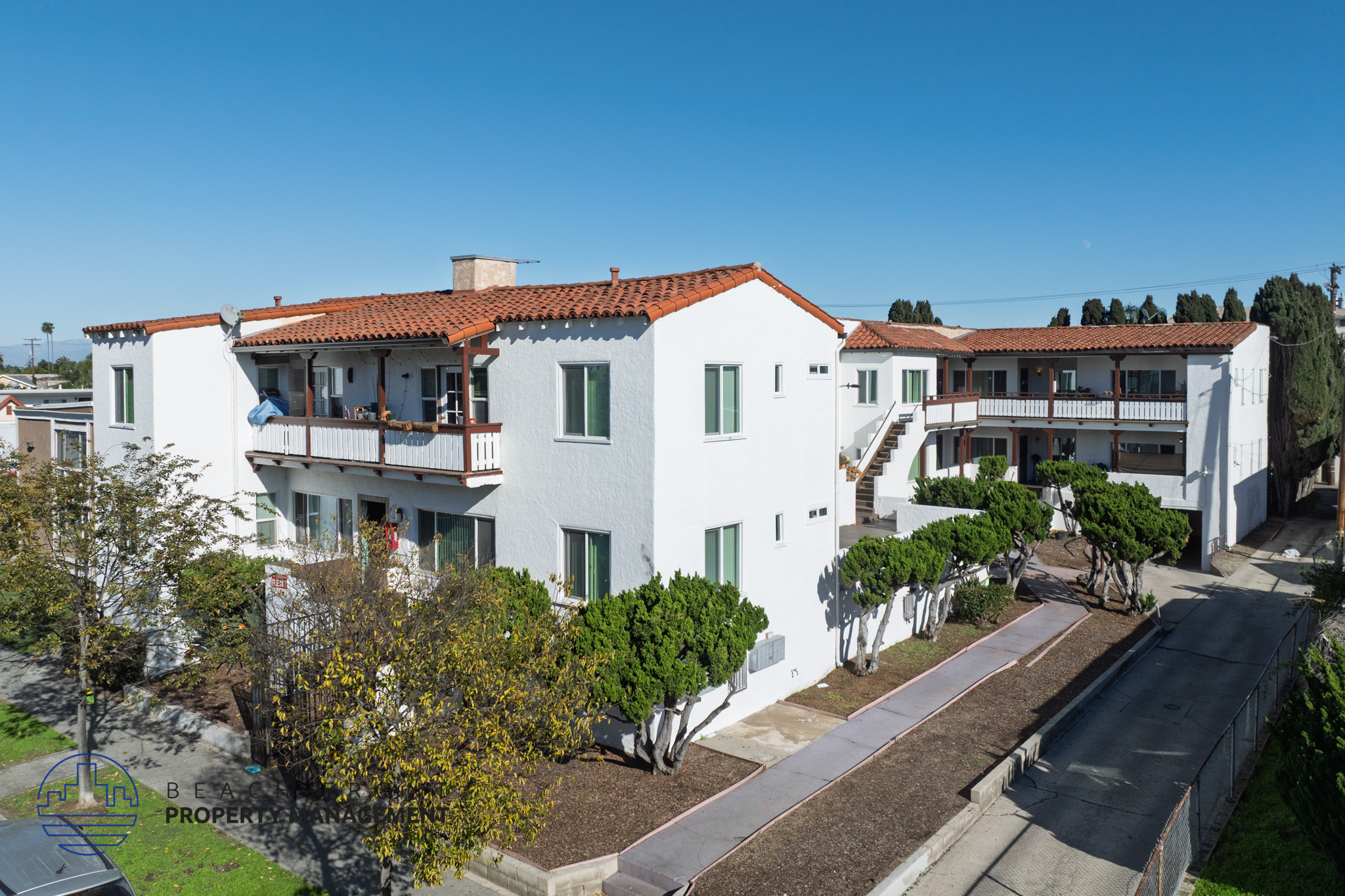 A white building with a red tile roof is surrounded by trees and shrubs.