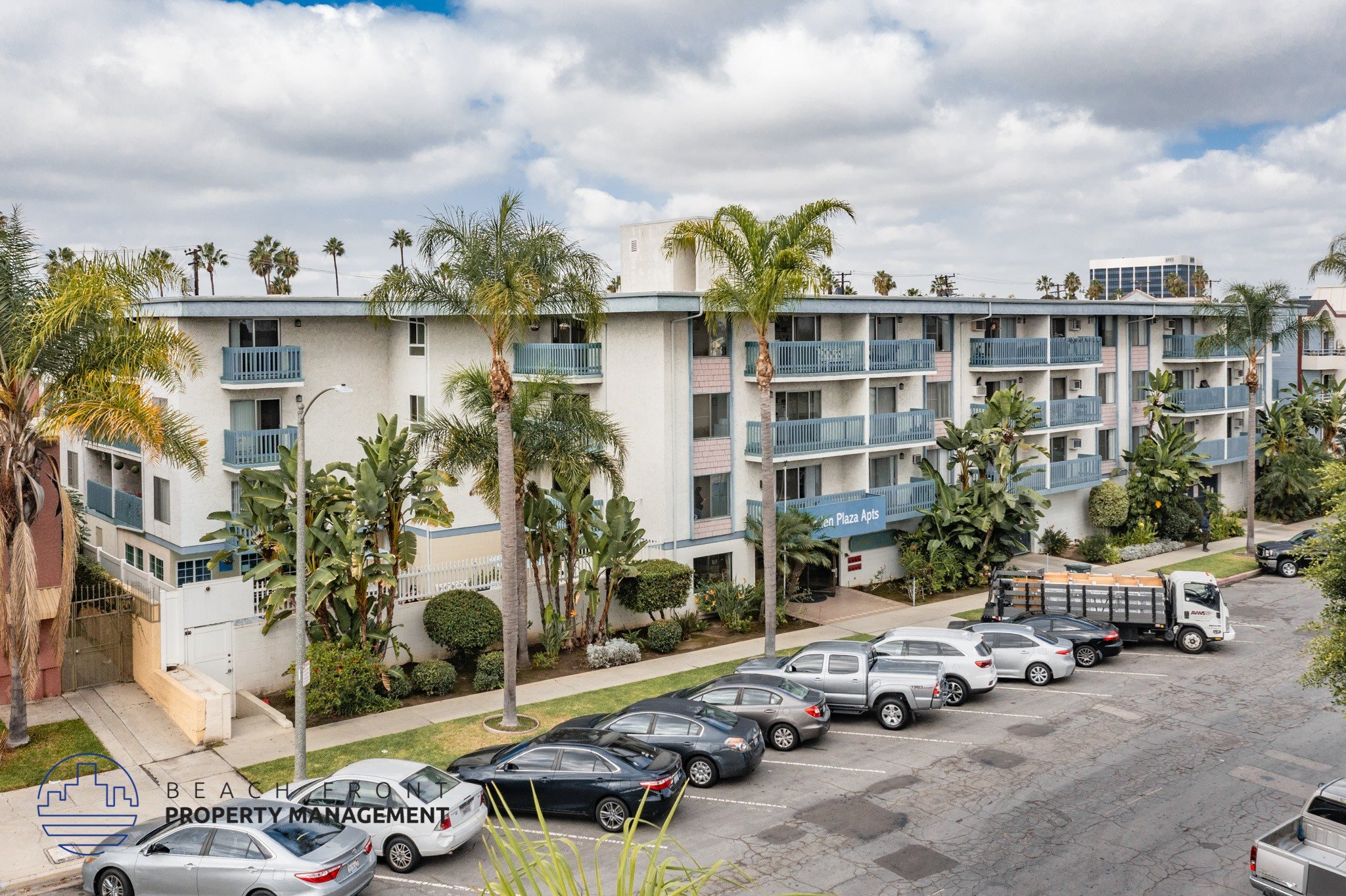 A parking lot with cars and a truck in front of a white apartment building with palm trees.
