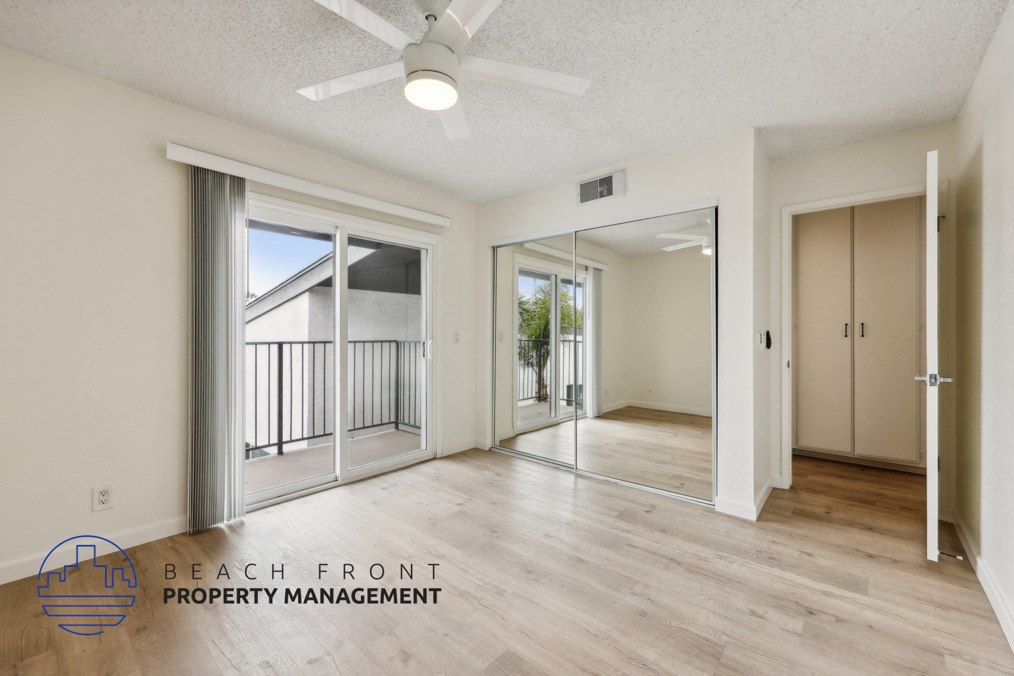A spacious room with a sliding glass door leading to a balcony, a ceiling fan, and a logo for Beach Front Property Management.