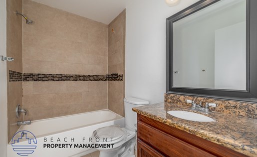 A bathroom with a beige tiled shower, a white toilet, a sink with a granite countertop, and a mirror above the sink.