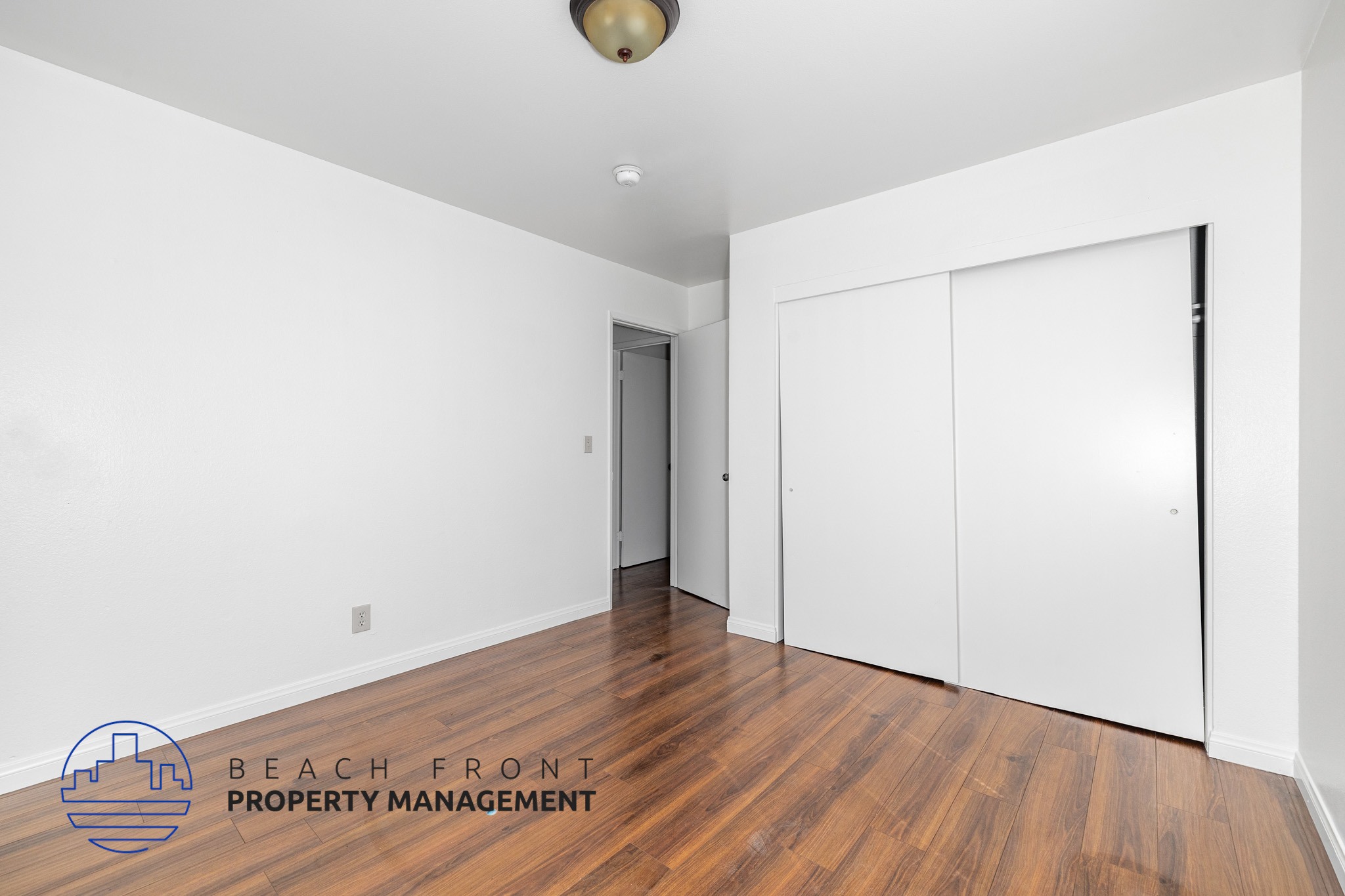 A room with a wooden floor and white walls, with a logo for Beach Front Property Management in the bottom left corner.