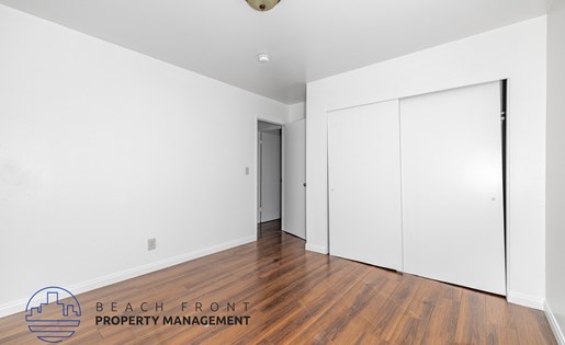A room with a wooden floor and white walls, with a logo for Beach Front Property Management in the bottom left corner.