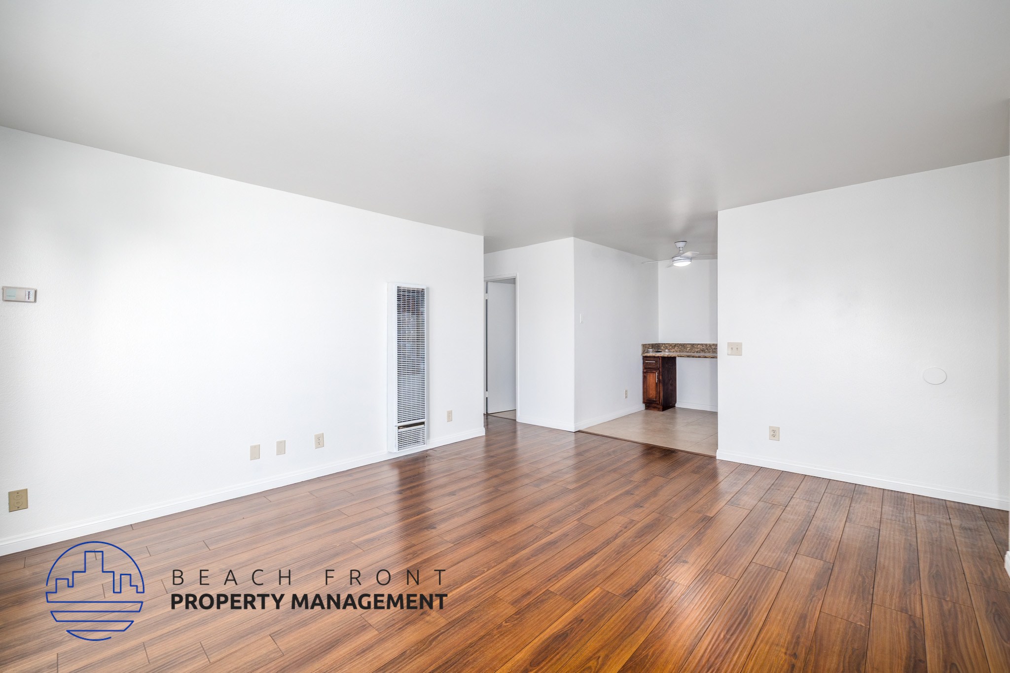 A spacious room with wooden flooring and white walls, with a logo for Beach Front Property Management in the bottom left corner.