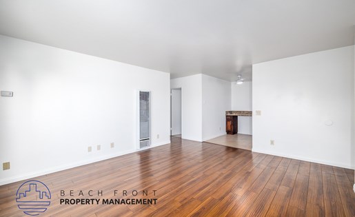 A spacious room with wooden flooring and white walls, with a logo for Beach Front Property Management in the bottom left corner.